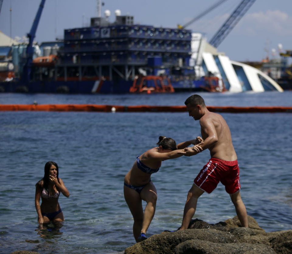 Sunbathers walk on rocks as the Costa Concordia cruiser is visible in background, in the Tuscan Island of Isola del Giglio, Monday, July 15, 2013. Salvage crews are working against time to right and remove the shipwrecked Costa Concordia cruise ship, which is steadily compressing down on itself from sheer weight onto its granite seabed perch off the Tuscan island of Giglio. Salvage master Nick Sloane said Monday that the Concordia has compressed some 3 meters (10 feet) since it came to rest on the rocks Jan. 13, 2012 after ramming a jagged reef during a stunt ordered by the captain that cost the lives of 32 people. (AP Photo/Gregorio Borgia)