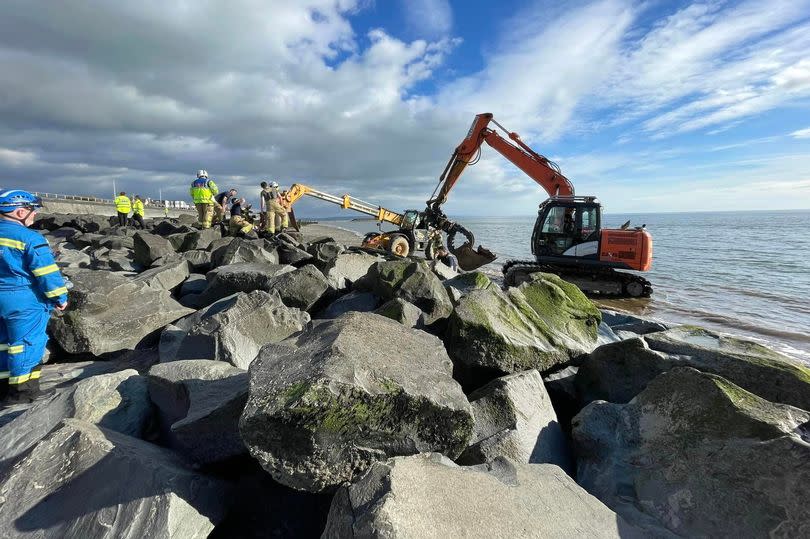 A digger operates on the shoreline as the advancing sea water begins to lap against the rocks