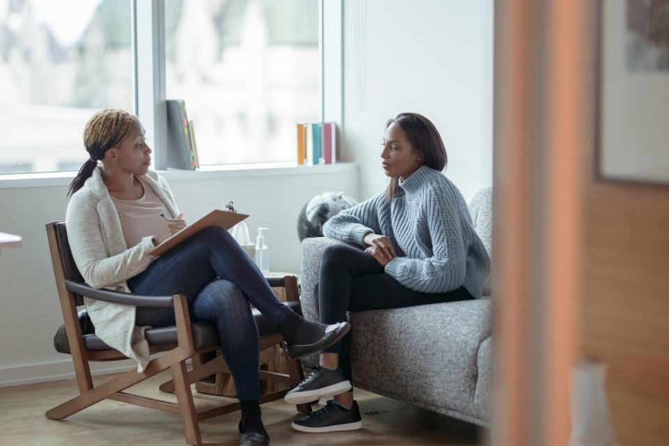 a therapist meets with her female client in her office the client is seated on a sofa with her arms across her body as she looks visibly nervous the therapist is seated in a chair in front of her as she talks about what to expect from the appointment and takes notes on her clipboard