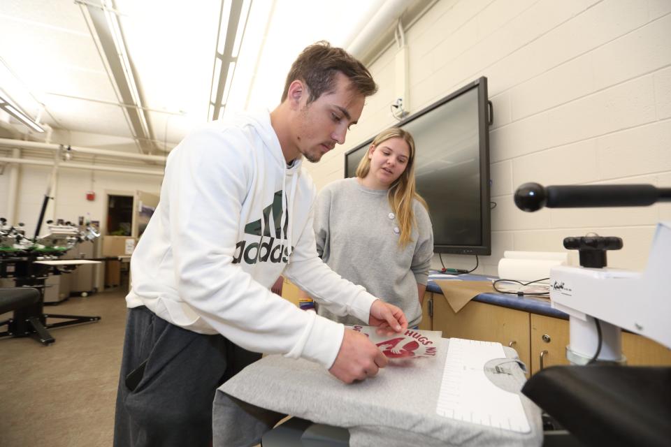 Canal Winchester High School senior Ethan McGlone (left) shows junior Elayna Griffith how to use an iron press to press a vinyl logo onto a shirt May 5.