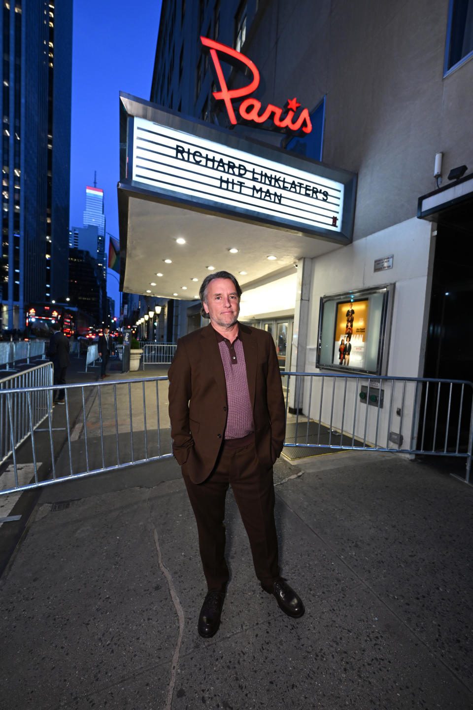 Richard Linklater in a brown suit stands outside a theater displaying a marquee that reads "Richard Linklater's Hit Man." City buildings and a lit sign in the background