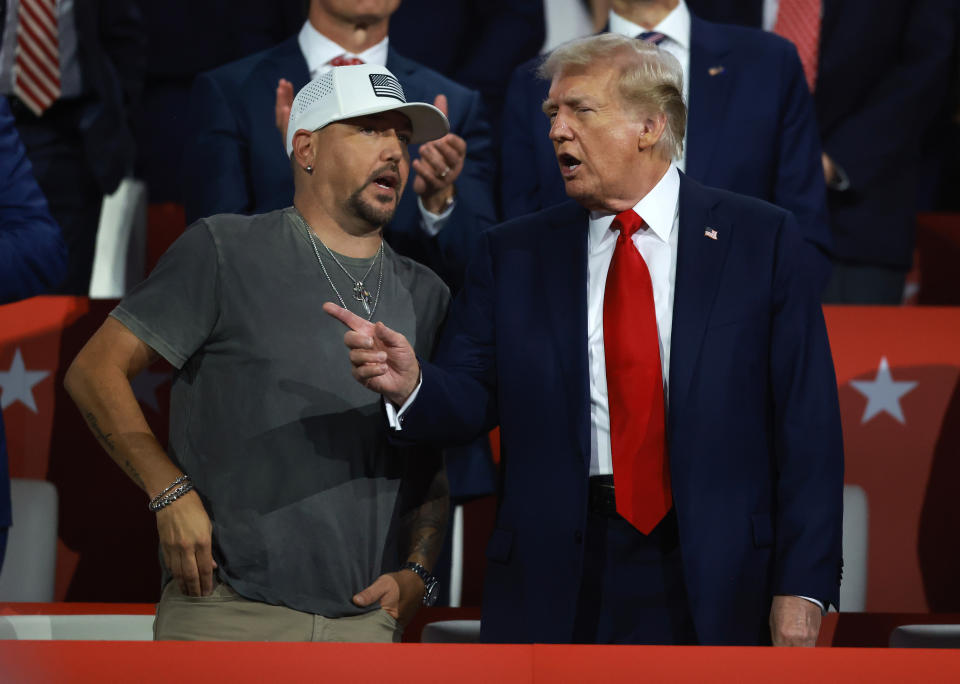 Jason Aldean und Donald Trump unterhalten sich angeregt bei einer öffentlichen Veranstaltung, wobei Jason lässig in T-Shirt und Mütze gekleidet ist und Trump einen Anzug trägt