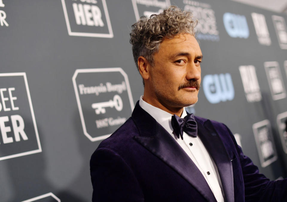 Taika Waititi in formal attire, including a bow tie and velvet jacket, poses on the red carpet in front of a backdrop displaying various logos