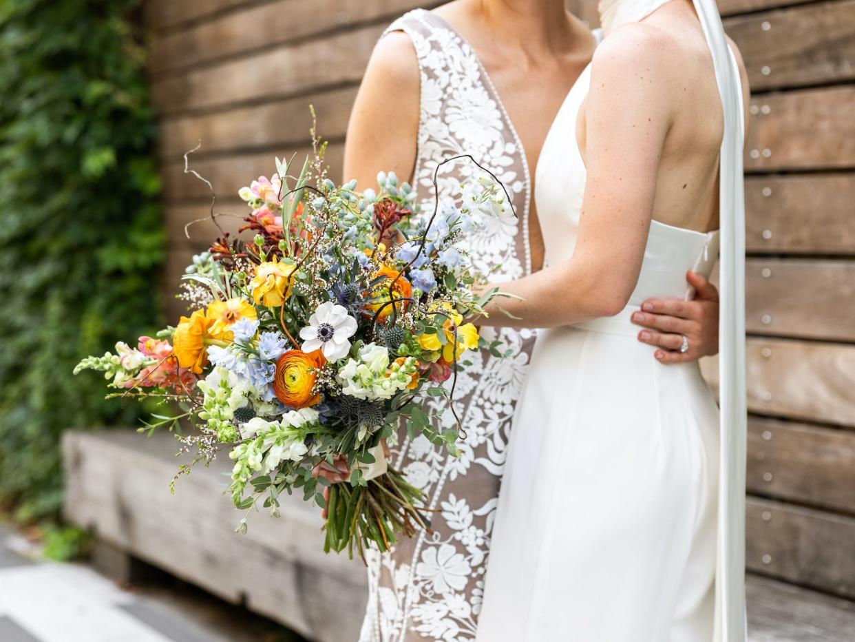 Two brides hug and hold a bouquet of colorful flowers.