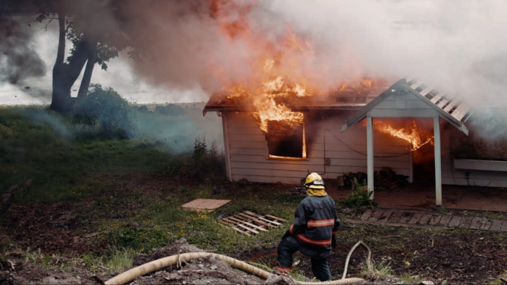 A woman firefighter works to move equipment, a burning unrecognizable home fully engulfed in fire and smoke in the background.