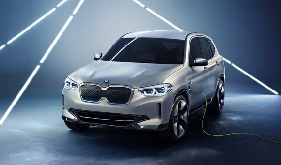 A prototype BMW iX3, a silver compact SUV, is shown plugged into a charger.