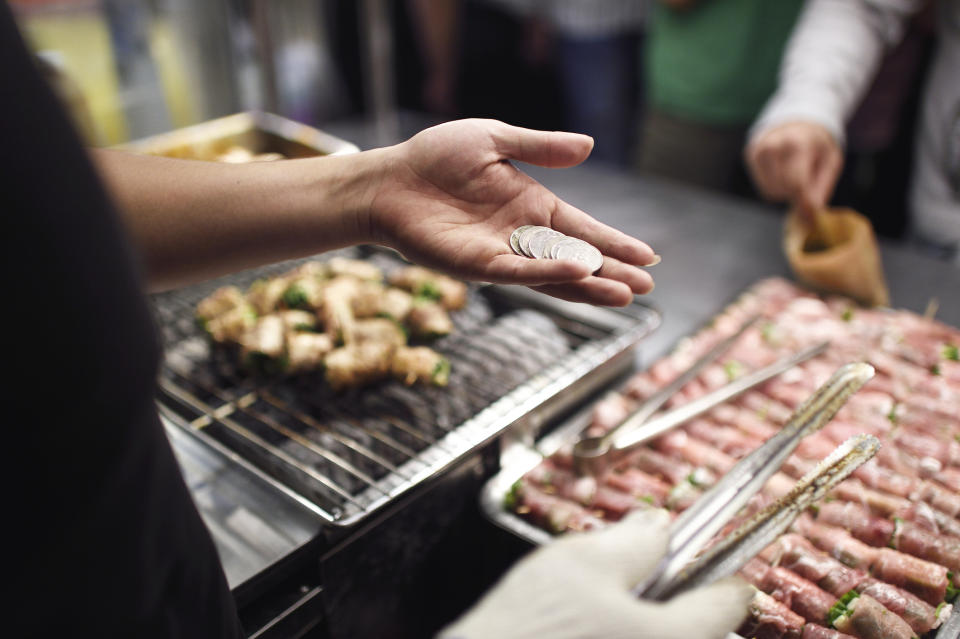 A person's hand holding coins near a food stall, with food items being cooked on a grill and another hand using tongs to pick up food