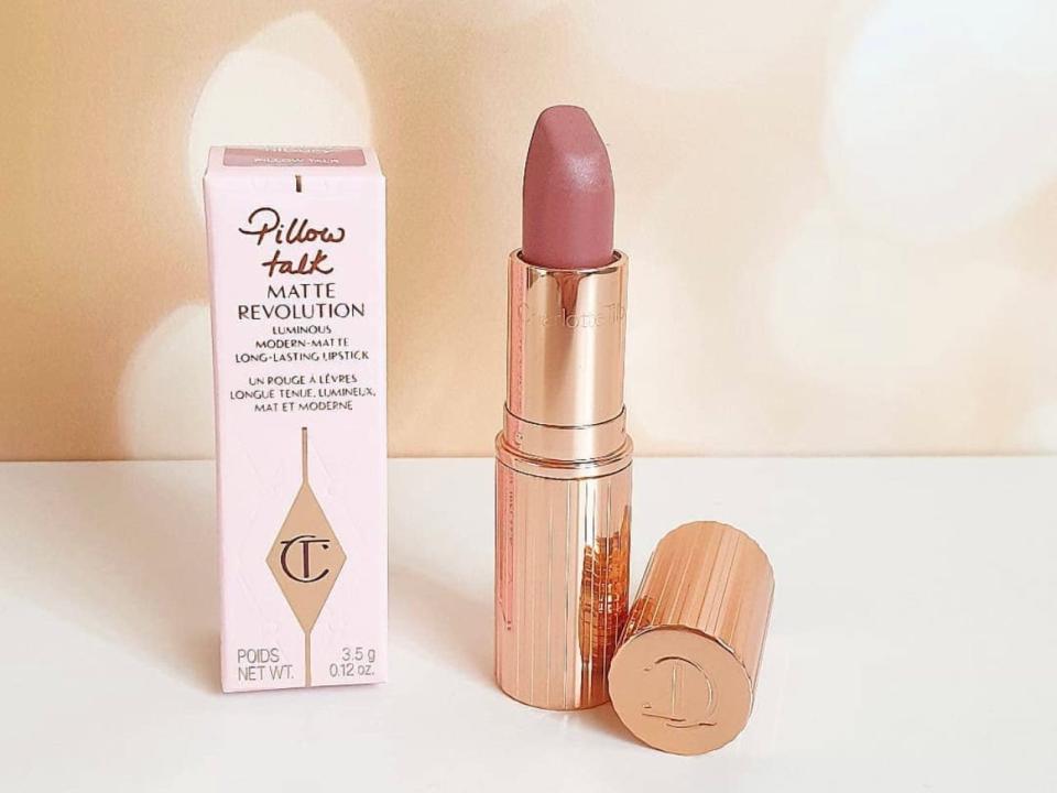 A pink box with loopy "Pillow Talk" text across the top next to a mauve-tone lipstick in a golden bullet on a white table and in front of a Champagne-colored backdrop