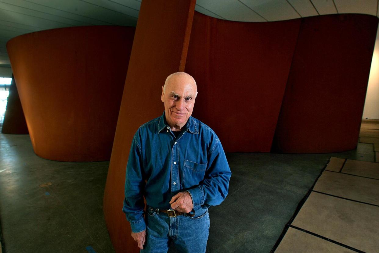 Richard Serra, in denim pants and shirt, stands in front of a curved sculpture with four expansive cavities.