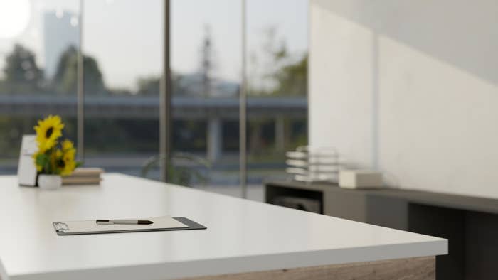 A modern office desk with a clipboard and pen, positioned in front of large windows showing an outdoor urban scene. Sunflowers and books are on the desk