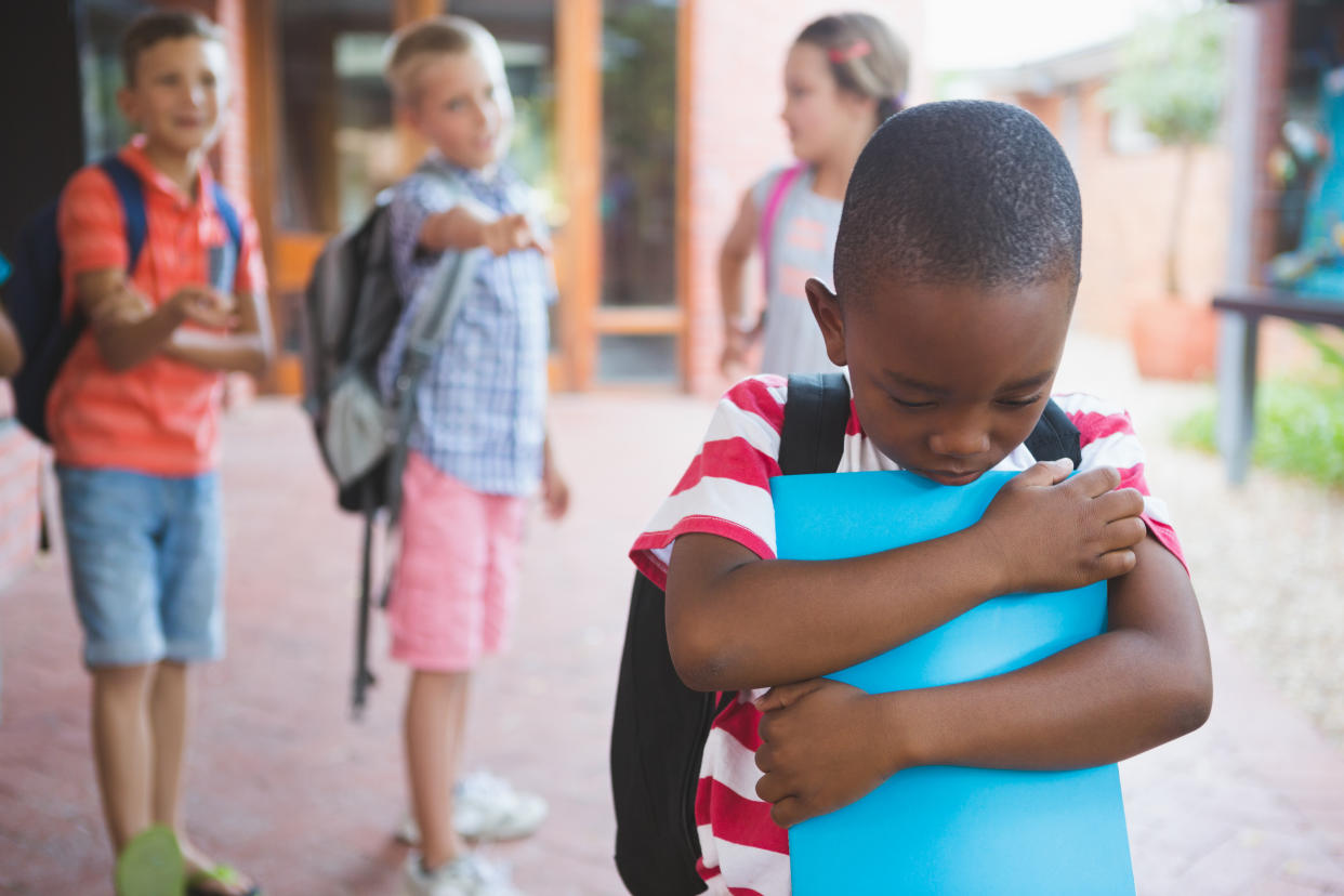 Three white elementary school children point at a Black boy, also wearing a backpack, who hugs a folder and looks despondently at the ground.