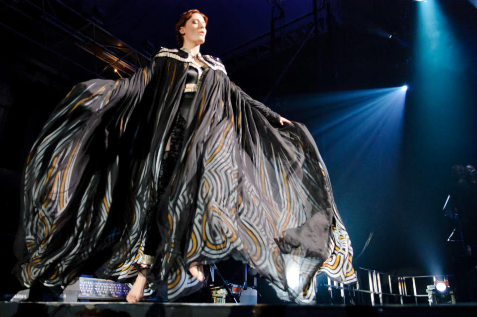 Florence Welch performs onstage in a black dress with matching cape. 