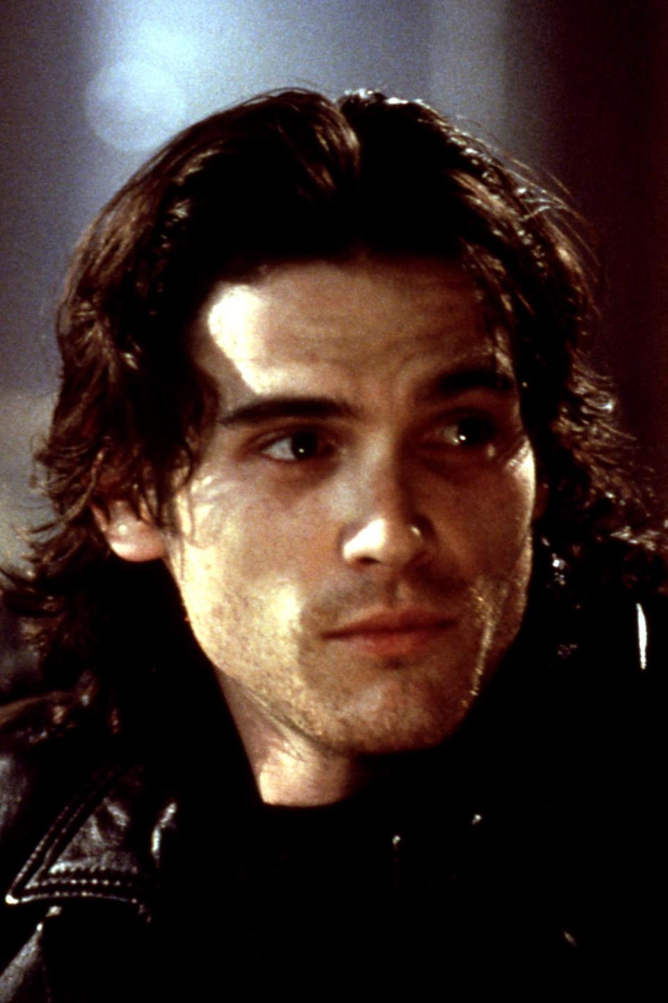 A young Billy Crudup with medium-length, wavy hair and wearing a leather jacket looks off-camera with a thoughtful expression