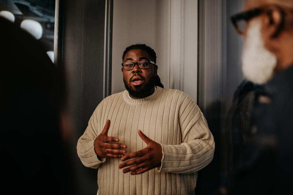 Man in sweater speaking during a meeting, gesturing with hands. Other attendees partially visible