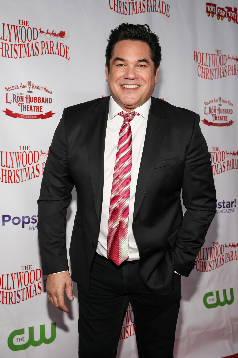 Dean Cain arrives on the red carpet at the Hollywood Christmas Parade, smiling for the cameras in a black suit and red tie.