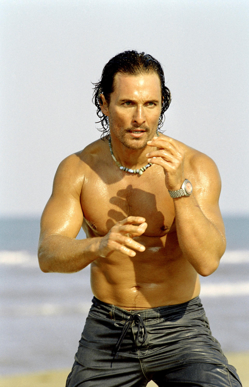 A shirtless man stands on a beach with his hands raised in a fighting stance, wearing a beaded necklace and watch