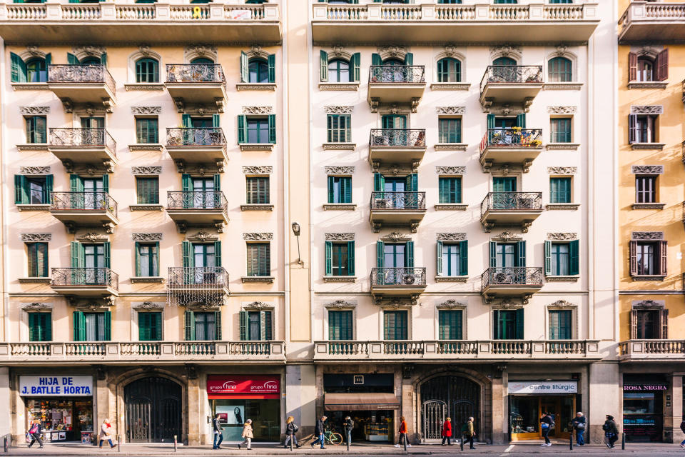 A street view of an apartment building with multiple balconies. Shops and cafes occupy the ground floor. Pedestrians walk along the sidewalk