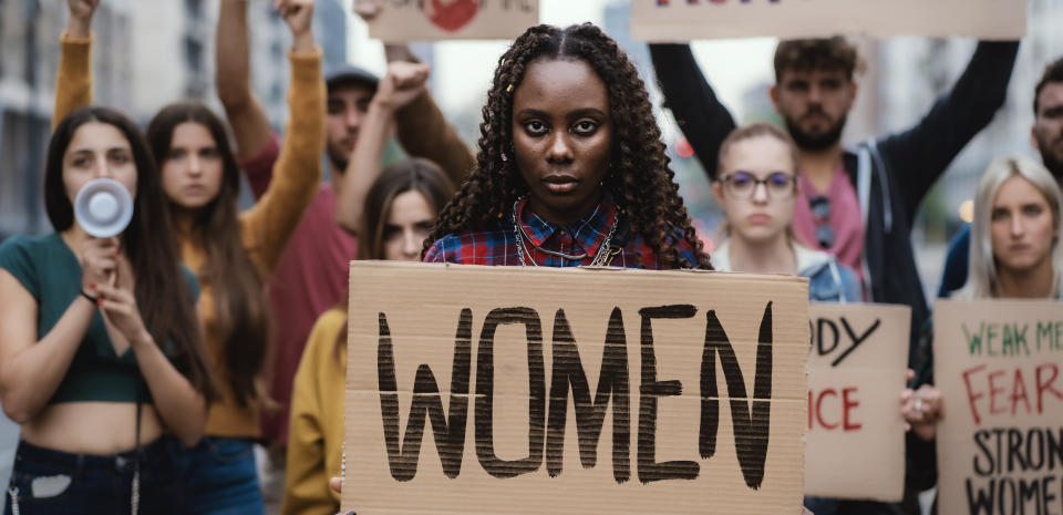 Multi-ethnic young protesters marching for women rights showing a signboard of Women. African woman in front holding the sign about woman empowerment.
