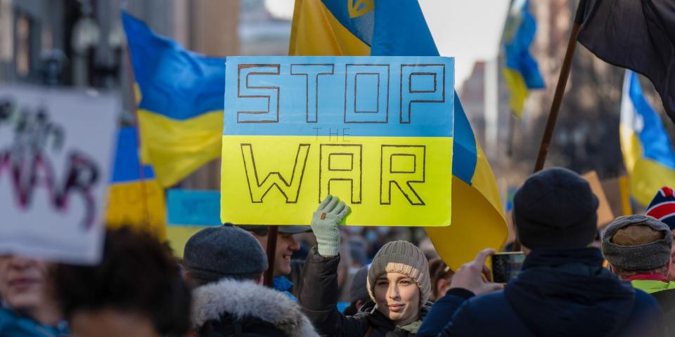 person in crowd of Ukraine flags holding blue-and-yellow sign that says "Stop War"