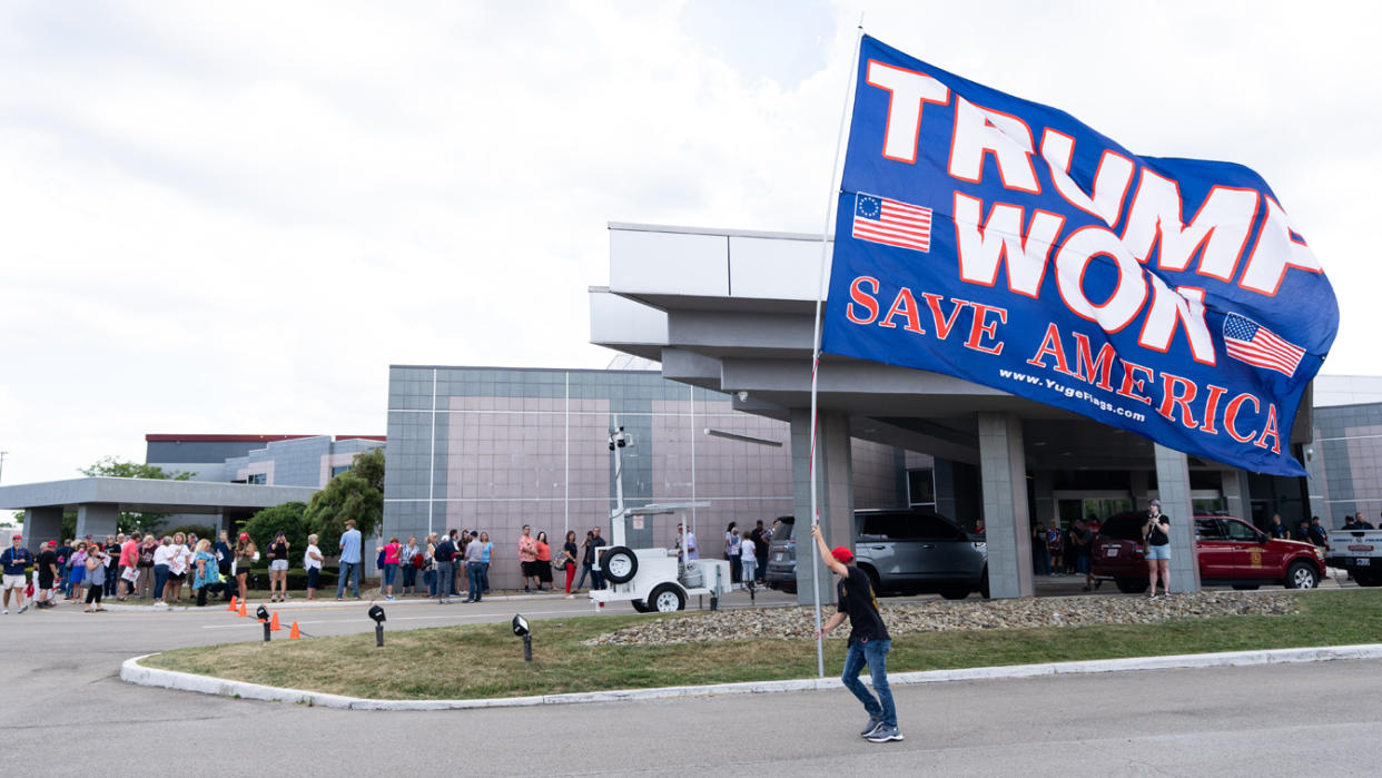 As people line up in the background to enter a rally, a man holds a huge flag that says: Trump Won Save America.