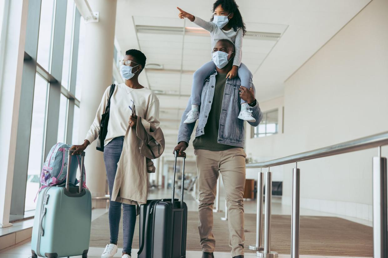 African father with young daughter on shoulder walking along wife with luggage at airport terminal. Family in pandemic with luggage at airport corridor looking away.