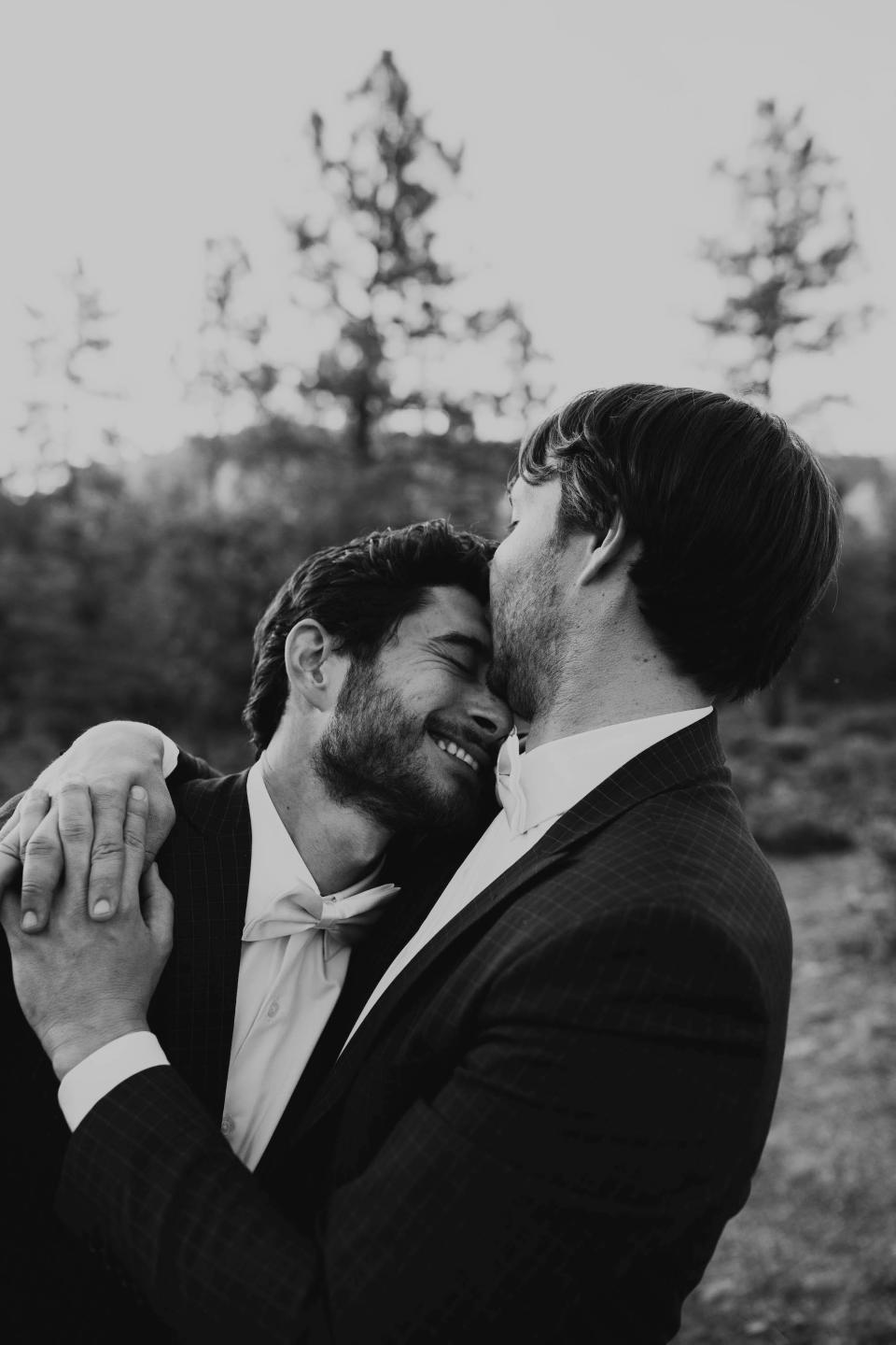 Two grooms embrace in a forest in their wedding attire.