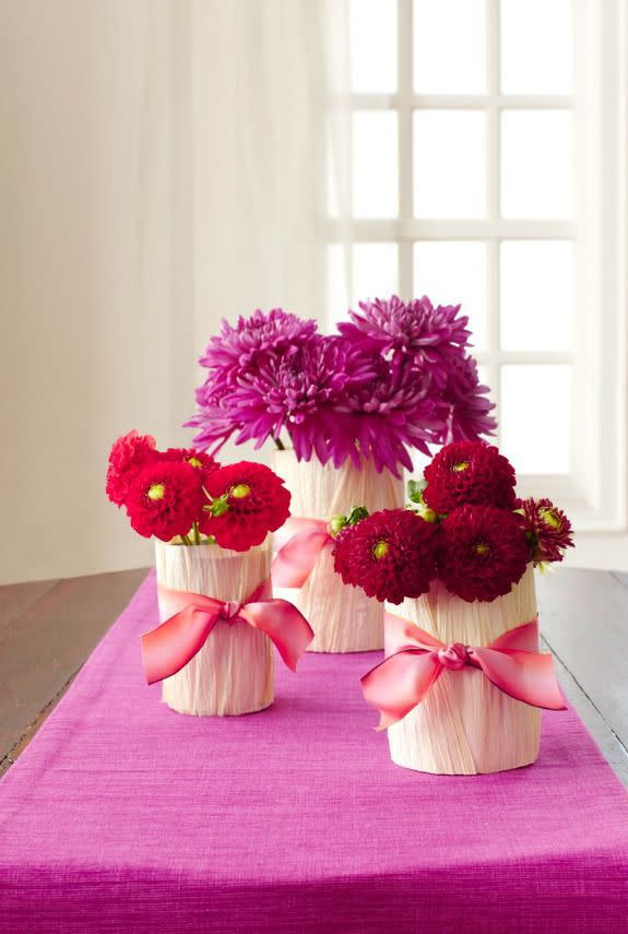 <p>Turn basic blossoms and kitchen cans into sweet arrangements for under $15. </p><p>Materials:</p><p> • Empty, cleaned food cans of varying sizes</p><p> • Pliers</p><p> • Hot-glue gun and glue sticks</p><p> • <a rel="nofollow noopener" href="http://www.amazon.com/El-Guapo-Corn-Husks-oz/dp/B0000GK6WQ/" target="_blank" data-ylk="slk:Dried corn husks;elm:context_link;itc:0;sec:content-canvas" class="link ">Dried corn husks</a></p><p>• Scissors</p><p> • Ombré ribbon </p><p> • 3 or more bunches of grocery store flowers (try spider mums and zinnias)</p><p> • Floral shears or kitchen scissors</p><p> • Water to fill cans</p><p>Directions:</p><p>1. Gather cans, making sure to press down any sharp edges with pliers.</p><p><strong>2.</strong> Apply a bead of hot glue vertically down the side of the first can and adhere a piece of corn husk. Hold it in place and let set. Continue until the entire can is covered. Trim the top and bottom edges with scissors until straight. Repeat on the remaining cans.</p><p><strong>3.</strong> Wrap a piece of ribbon around each can and knot. Trim edges to prevent fraying.</p><p><strong>4.</strong> Choose a flower bunch and a can (for a cohesive look, stick to one color of flower for each arrangement). Hold the flower stems up to the side of the can to gauge how tall they should be (aim for a height just slightly higher than the top of the can). Snip ends with shears or scissors at an angle to allow them to soak up more water.</p><p><strong>5.</strong> Fill each can with cool tap water and pour in the food packet that came with the bouquet. Place flowers in the can and fluff slightly until they sit comfortably. Repeat with other flowers until each can is filled. Arrange along the middle of the table.</p>