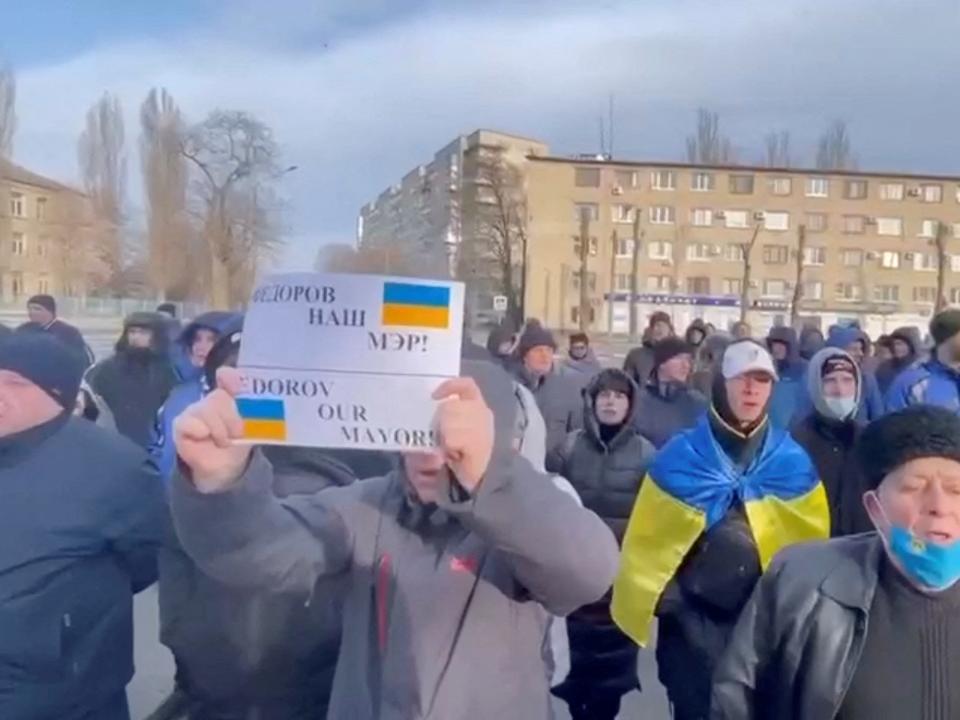 A crowd of Ukrainian men stand outside in Melitopol holding up signs in protest of the abduction of their mayor.