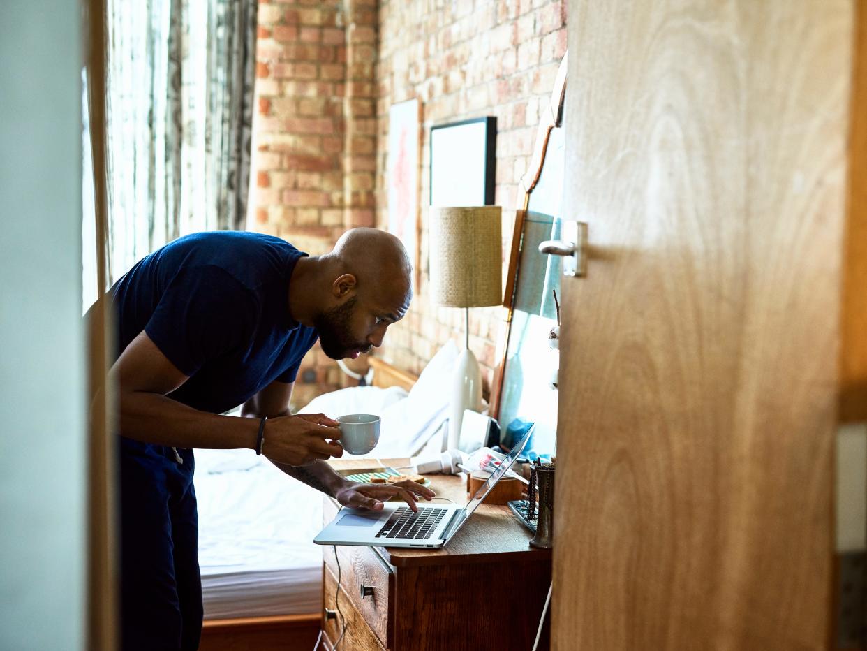 Man with coffee checking emails on laptop in bedroom