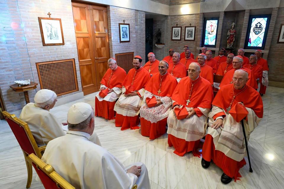 Pope Francis and Pope Emeritus Benedict XVI in white cassocks sit in front of a group of seated cardinals in red and white.