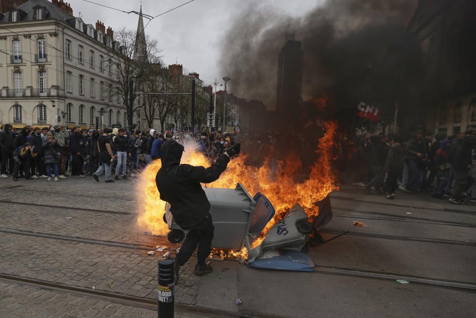 A protester stands next to a burning waste container during a rally in Nantes, western France, Thursday, march 23, 2023. French unions are holding their first mass demonstrations Thursday since President Emmanuel Macron enflamed public anger by forcing a higher retirement age through parliament without a vote. (AP Photo/Jeremias Gonzalez)