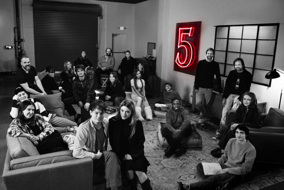 the cast of stranger things with matt and ross duffer, shown posing together on couches and rugs in black and white with a lit up number five in the corner of the room
