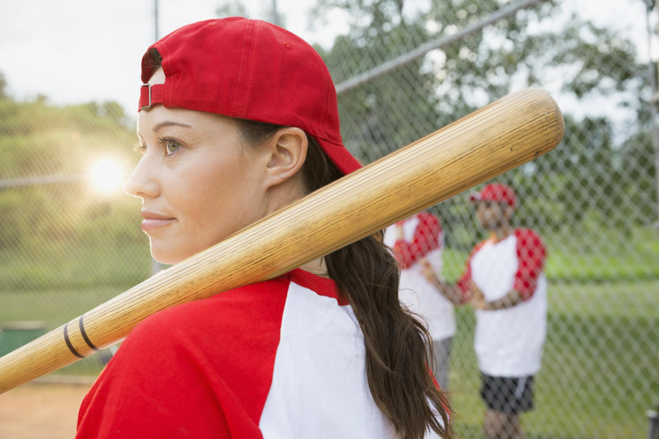 An image of a woman playing baseball. She's wearing a red cap backwards and has the bat resting on her shoulder.