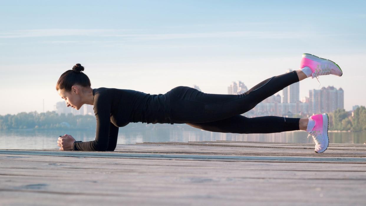  Woman performing a forearm plank outdoors by the water with left leg lifted in the air. 