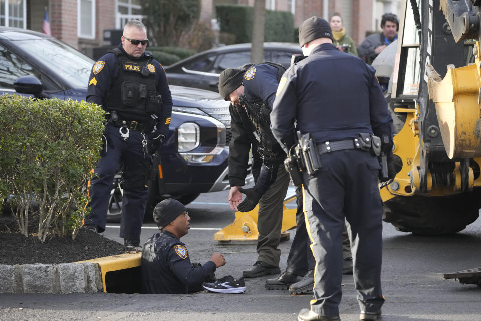 Several police stand near a drainage grate as one searches inside it near Dwumfour’s home.