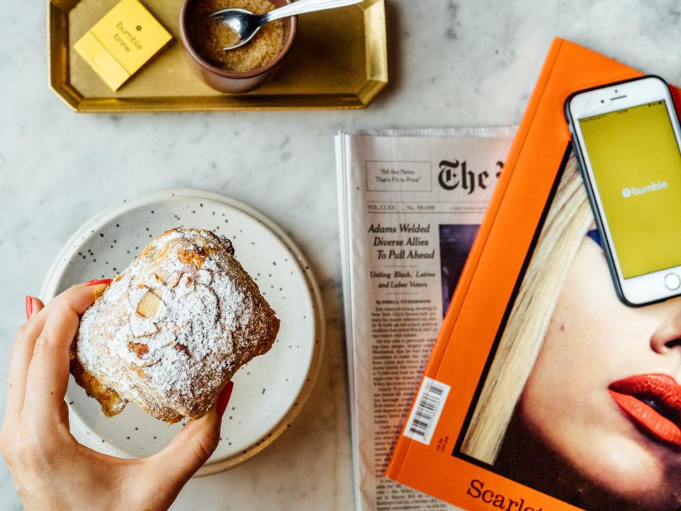 A hand holding a pastry with a newspaper, book, and iPhone showing the Bumble app off to the side.