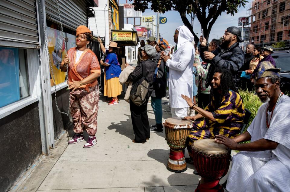 Percussionists play bongos on a sidewalk as a crowd watches two women roll up the metal security doors of a building