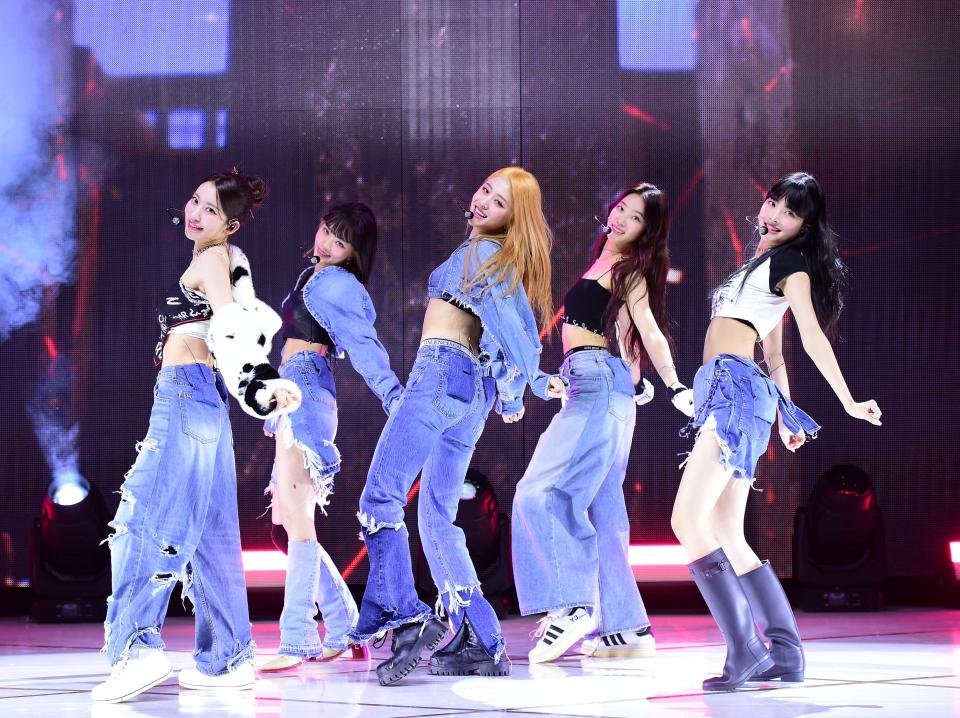 fiven young women in outfits comprised of black, white, and denim, posing onstage while they dance. their left feet have the heel up and their arms are extended behind them as they look over their left shoulder