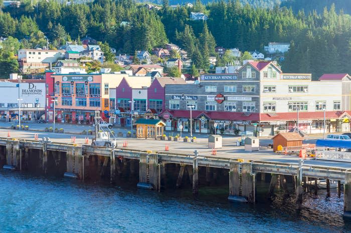 Promenade of colorful shops and buildings along a waterfront in a scenic town with forested hills in the background