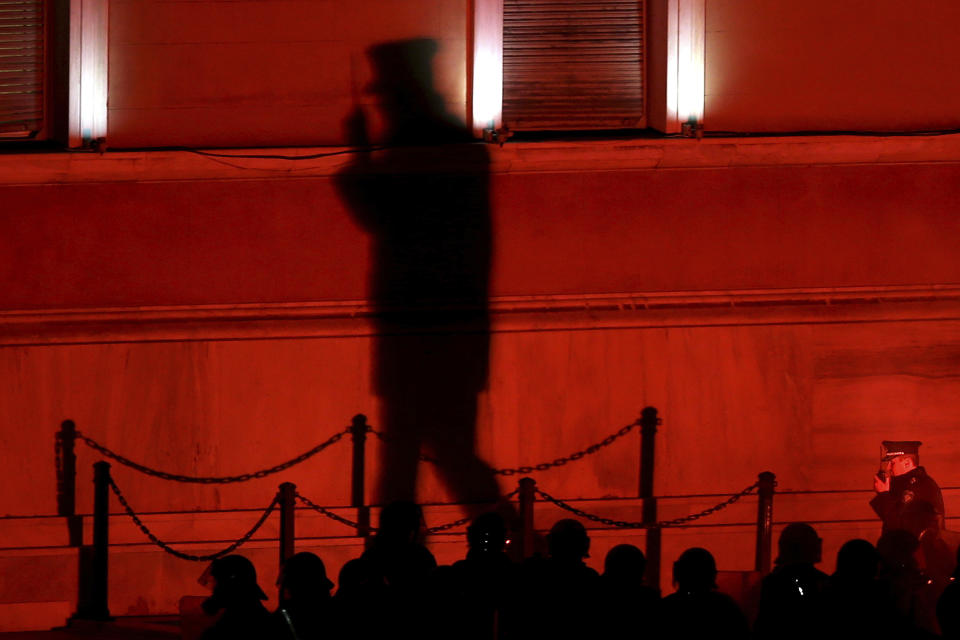 A shadow of a police officer on a wall of parliament use his radio and riot police officers, in silhouettes, stand guard as opponents of Prespa Agreement throw a flare during a rally outside the Greek Parliament in Athens, Thursday, Jan. 24, 2019. Greek lawmakers are debating a historic agreement aimed at normalizing relations with Macedonia in a stormy parliamentary session scheduled to culminate in Friday vote. (AP Photo/Petros Giannakouris)