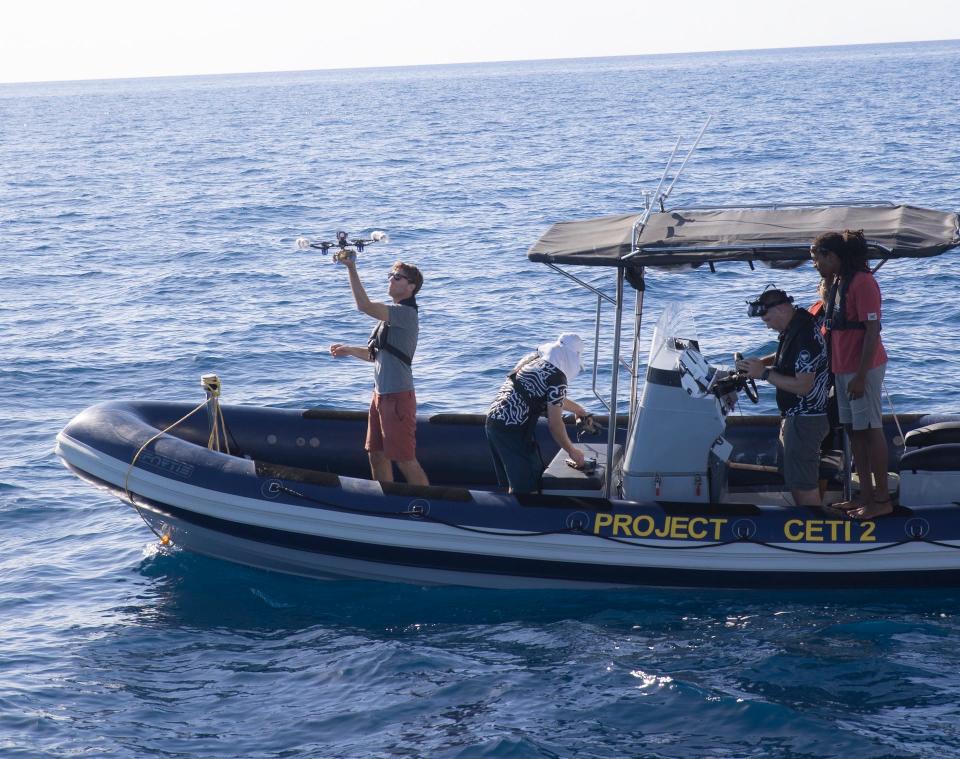 A group of four people on a small boat on the open ocean. One person is holding a drone at the front of the boat.