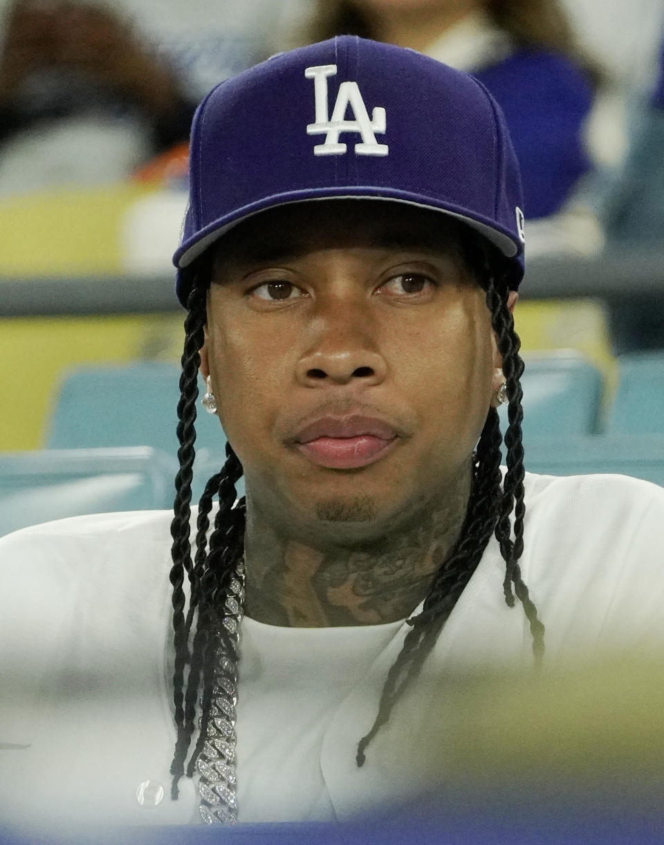 Tyga at a sporting event, wearing a Los Angeles baseball cap and a chain necklace, looking ahead