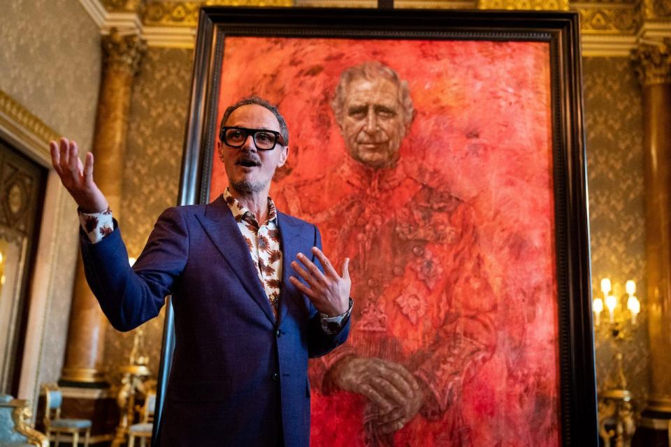 British artist Jonathan Yeo, wearing a blue suit and dark-rimmed glasses, stands in front of the portrait he painted of King Charles III at Buckingham Palace.