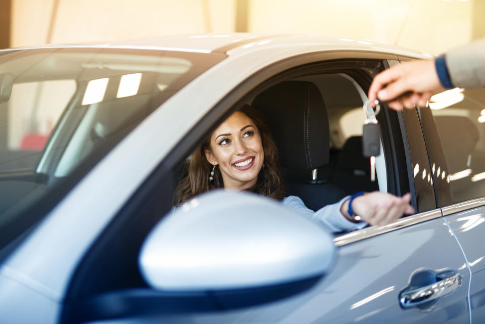 An attractive brunette woman sitting in her brand new car and taking keys from vehicle dealer.