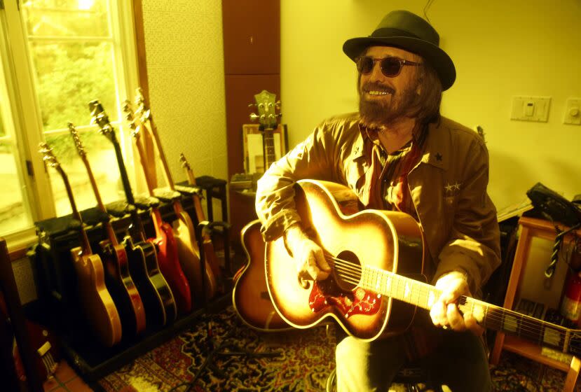 Tom Petty, dressed in a jacket, sunglasses and fedora, smiles and holds a guitar while seated in his home.