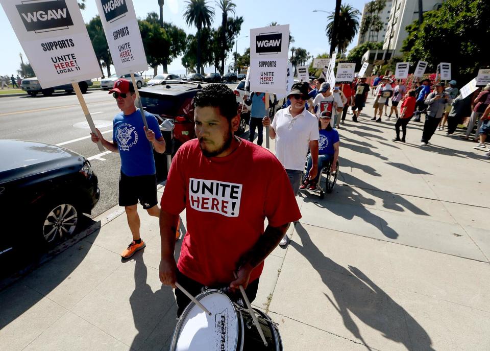 People hold signs and picket, one in a red "Unite Here" shirt, on a sidewalk with palm trees in the background