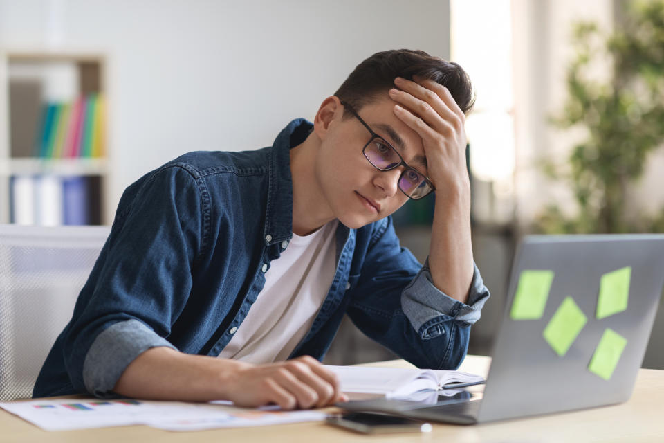 bored and vacant looking young worker staring at his laptop with his hand to his forehead