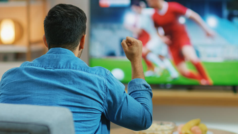 At Home Sports Fan Watches Important Soccer Match on TV, He Aggressively Clenches the Fist, Cheering for His Team. Cozy Living Room with Snacks and Drinks on the Table.