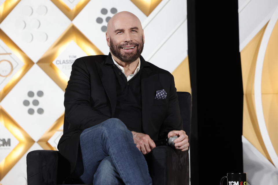 John Travolta sits onstage during an event, wearing a dark suit jacket, black shirt, and patterned pocket square, smiling and holding a microphone