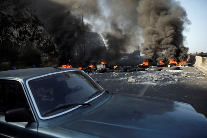 A man sleeps in a car next to burning tires barricading the highway during ongoing anti-government protests at Nahr El Kalb